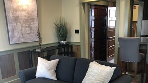 Uma sala de estar com um sofá cinza escuro, duas almofadas brancas, uma mesinha de centro de madeira com um vaso de flores brancas, um tapete estampado e uma pequena mesa redonda.