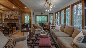 A cozy living room with a stone fireplace, wooden ceiling, and large windows offering a view of the forest.