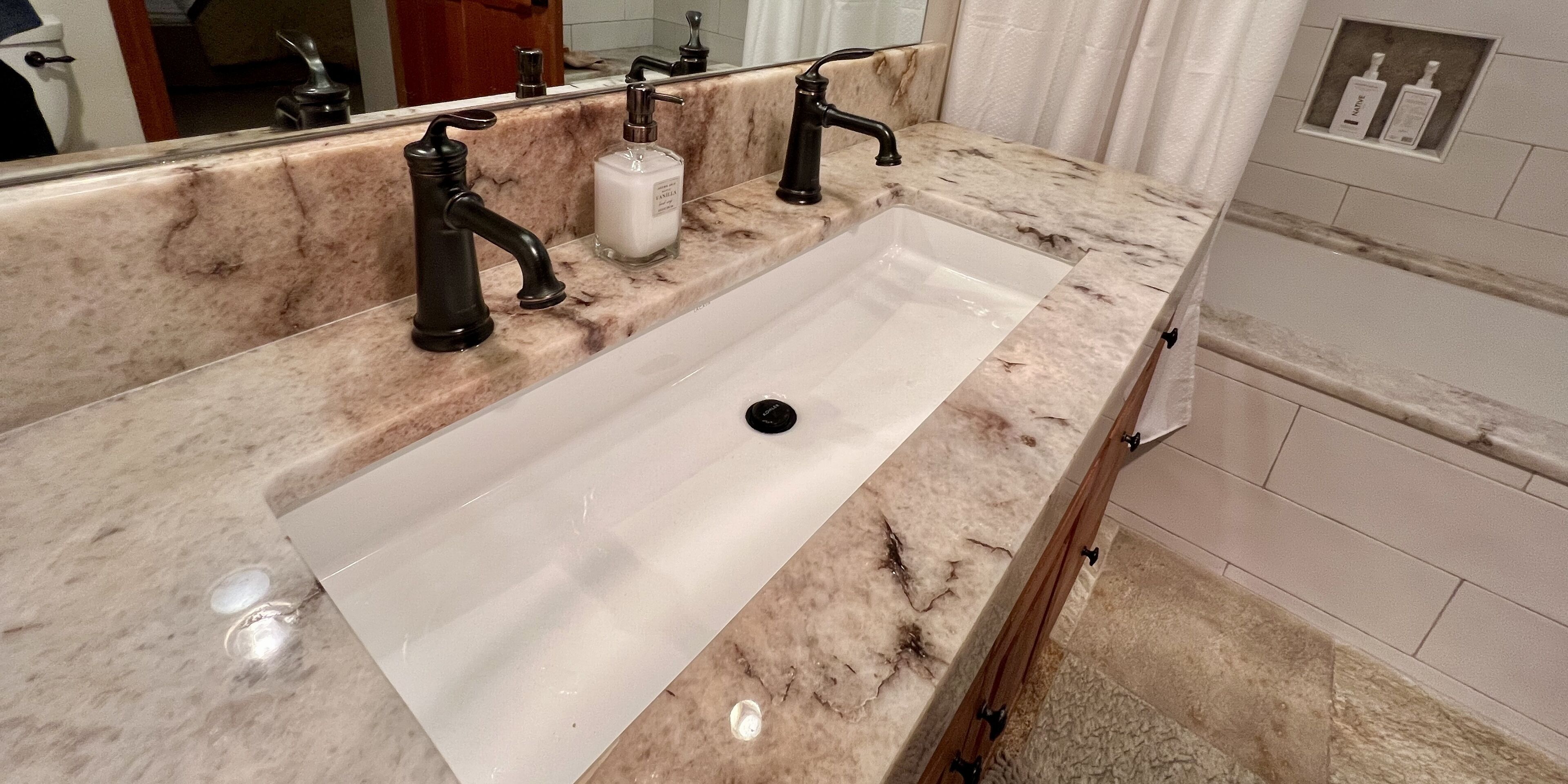 Une salle de bain avec un comptoir en marbre, un lavabo blanc et deux robinets noirs.