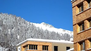 A building with a brick facade and large windows, set against a backdrop of snow-covered trees and a mountain. - Popular and centrally located residential building in St. (St. Moritz)