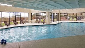 Una piscina interior con paredes de vidrio transparente, suelo de baldosas y varias sillas de descanso.