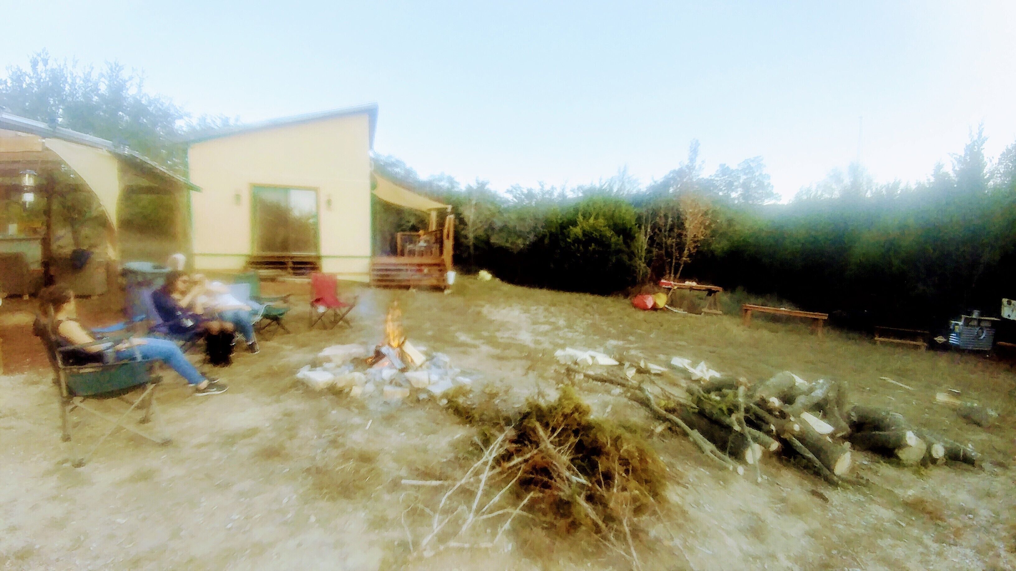 A rustic outdoor setting with a small building, a fire pit, and a few people sitting on chairs.