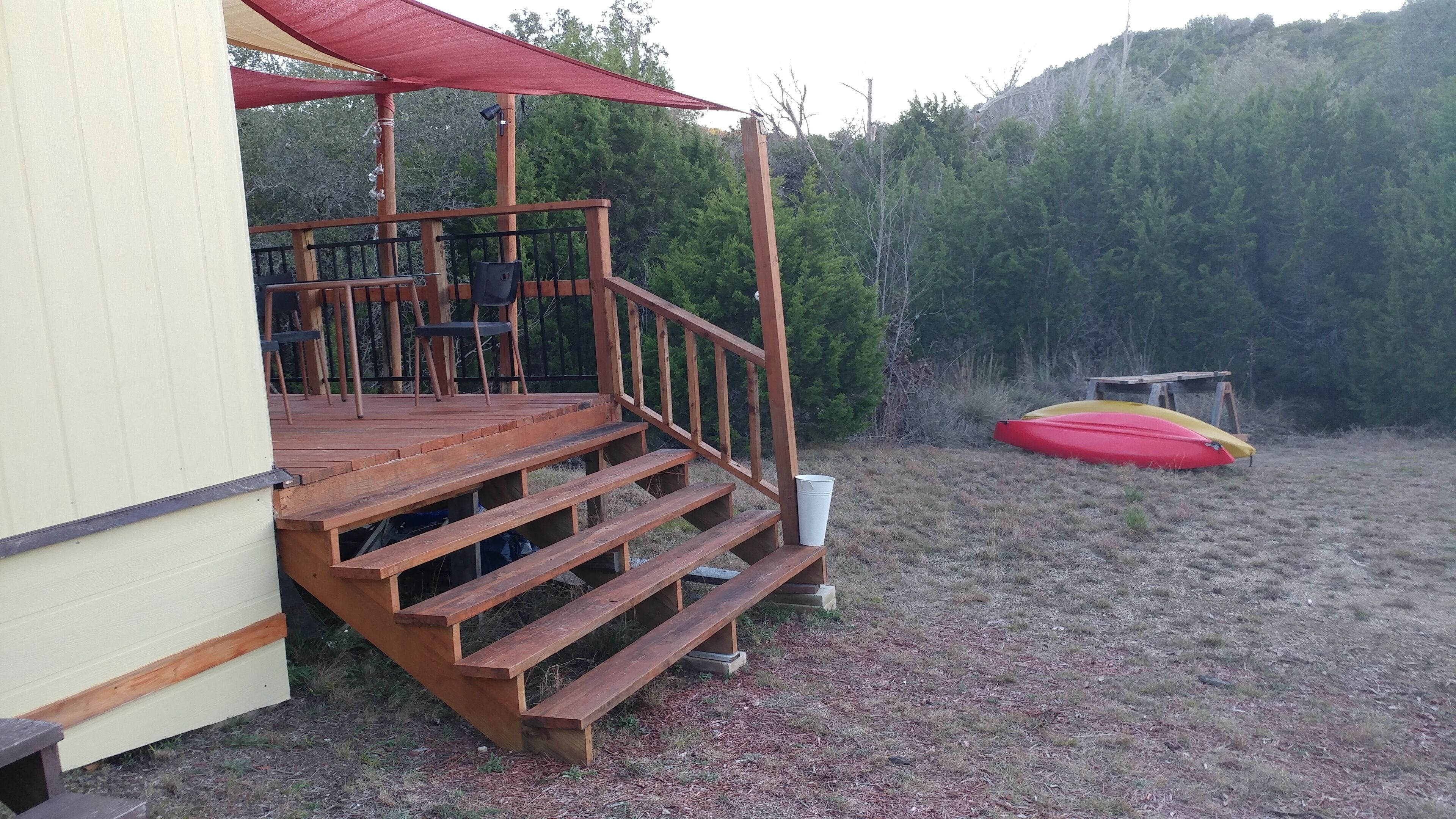 A wooden deck with steps leading to a covered area, a kayak, and a forested hillside in the background.