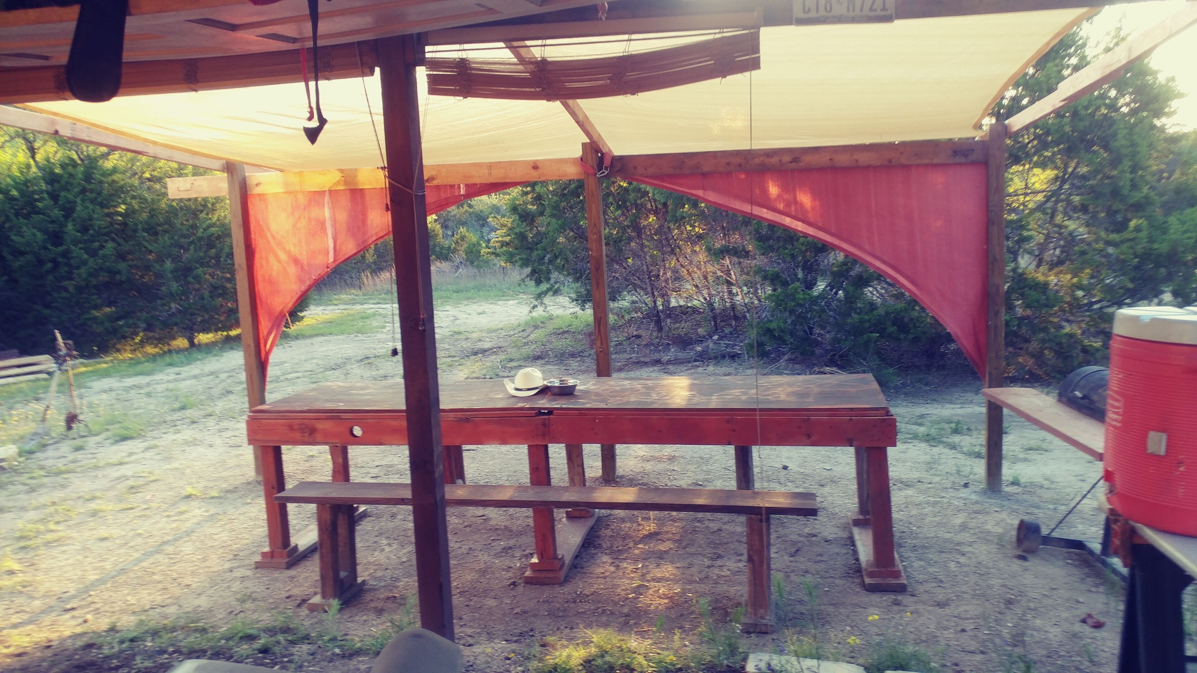 A wooden picnic table under a covered area with a bench, surrounded by trees and a gravel path.