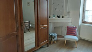 A room with a wooden wardrobe, a small round table, a chair with a striped cushion, a fireplace, and a window with lace curtains. - Pretty Potters' House (Accolay)