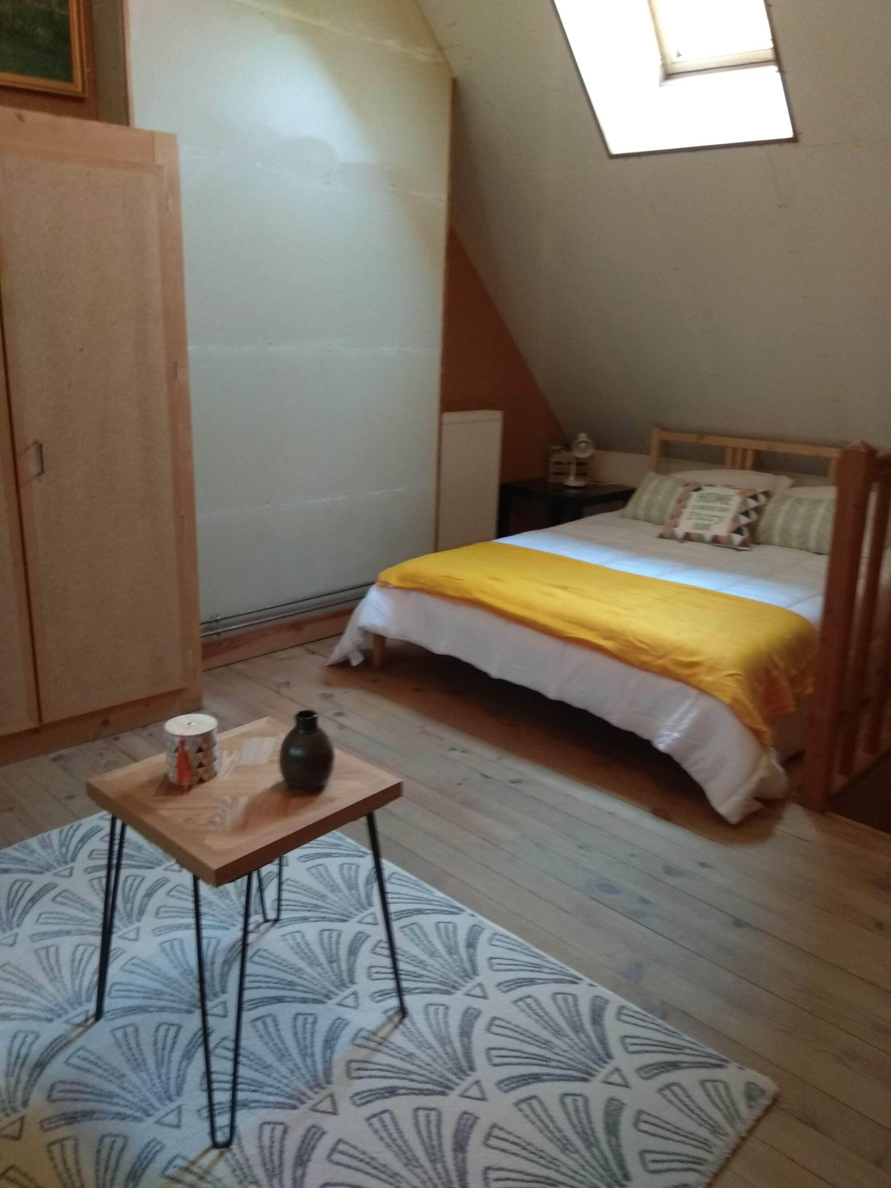 Quarto com cama, uma mesinha com vela e um vaso, guarda-roupa de madeira e teto inclinado com claraboia.