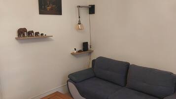 A living room with a grey sofa, wooden shelves with decorative items, a table, and a wall-mounted lamp.
