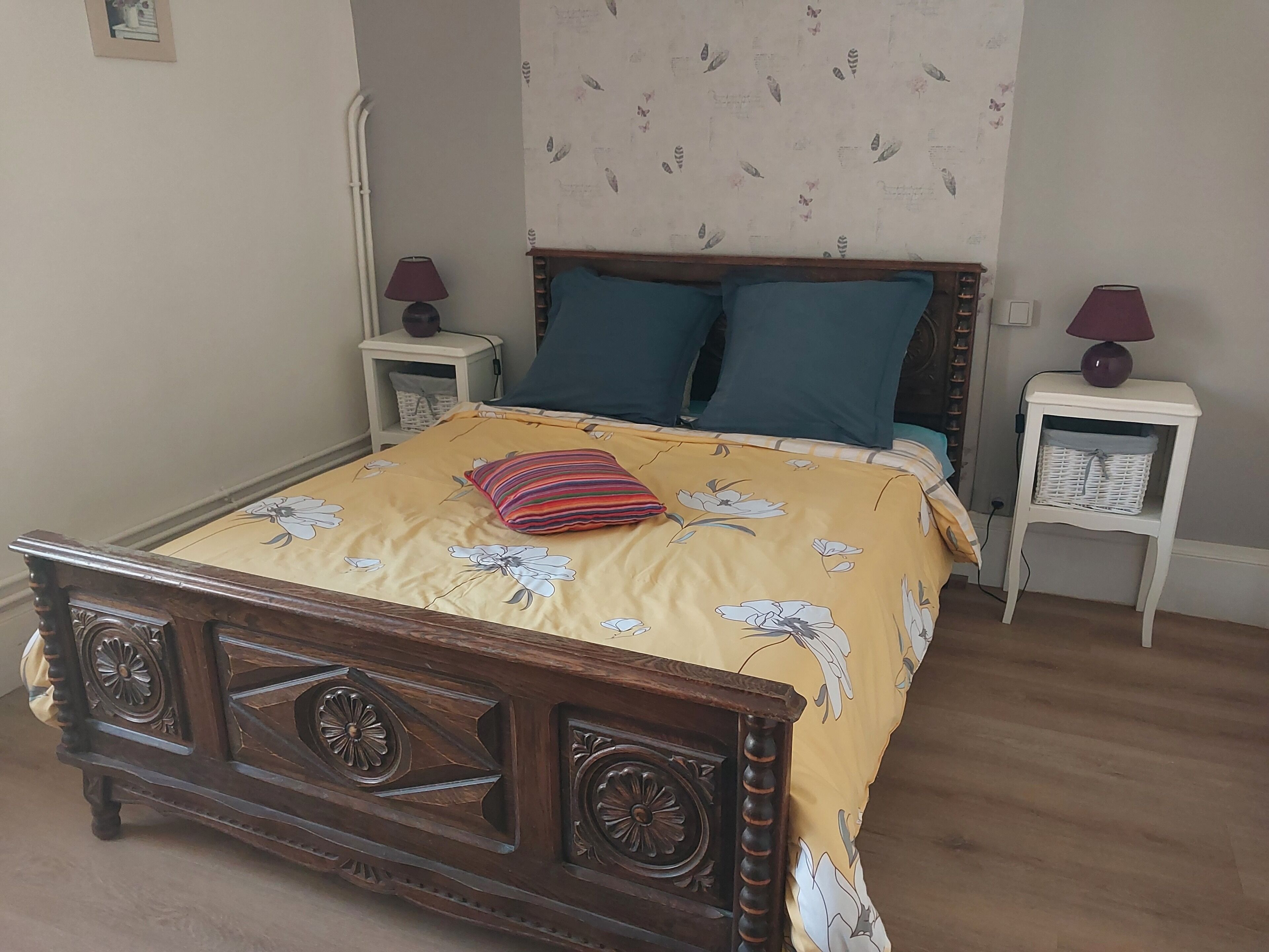 Een slaapkamer met een houten bed, een beddegoed met bloemenprint, twee kussens, een nachtkastje met een lamp en een wand met een bladmotief.