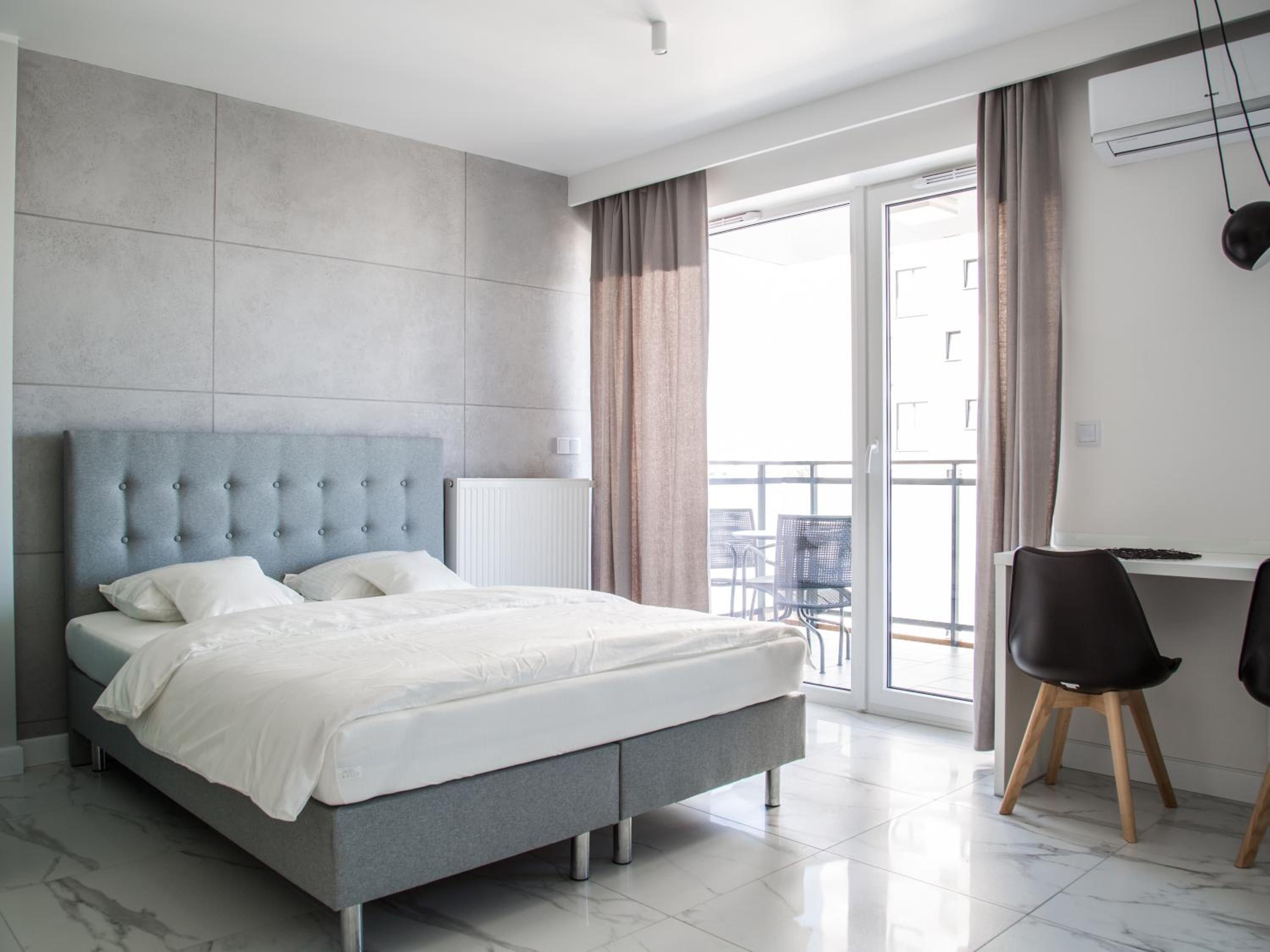 A modern bedroom with a grey bed, white bedding, a grey headboard, and a view of the outdoors through a sliding glass door.