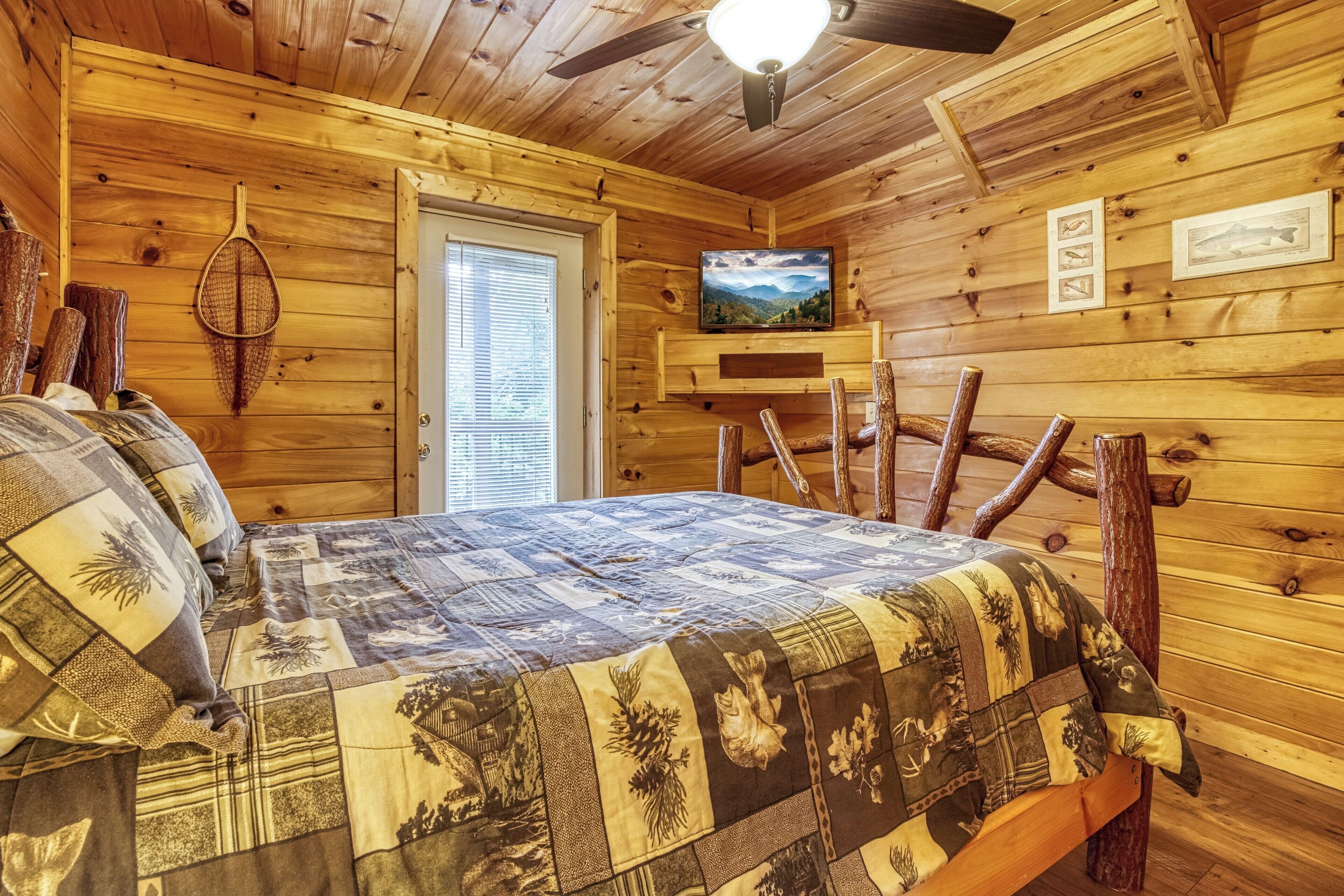 A wooden cabin bedroom with a bed, a ceiling fan, a television, and a door.