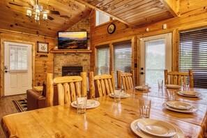 Un intérieur en bois avec une table à manger pour quatre personnes, une cheminée en pierre et un téléviseur fixé au mur.