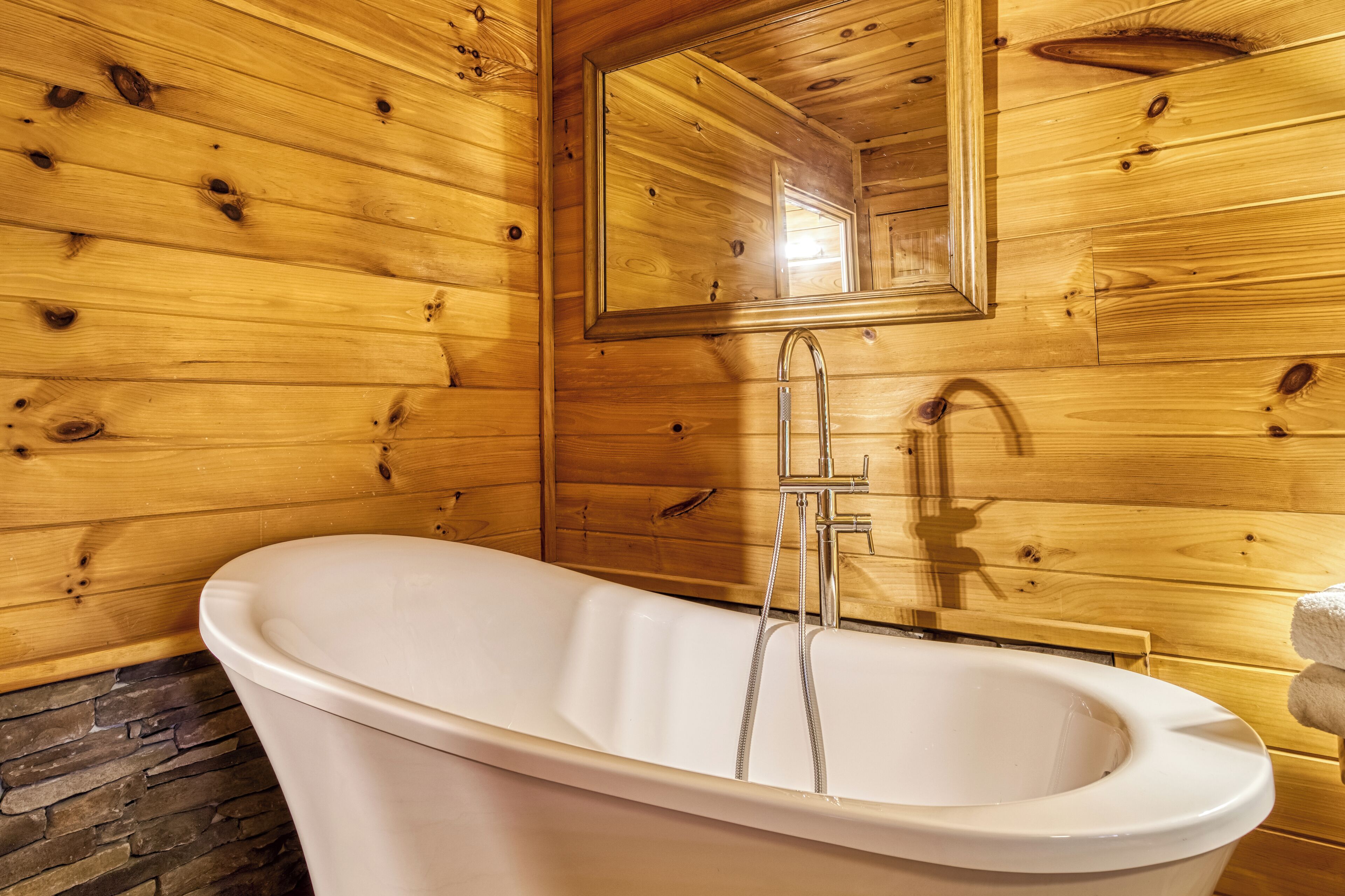 A wooden cabin bathroom with a freestanding bathtub, a mirror, and a stone wall.