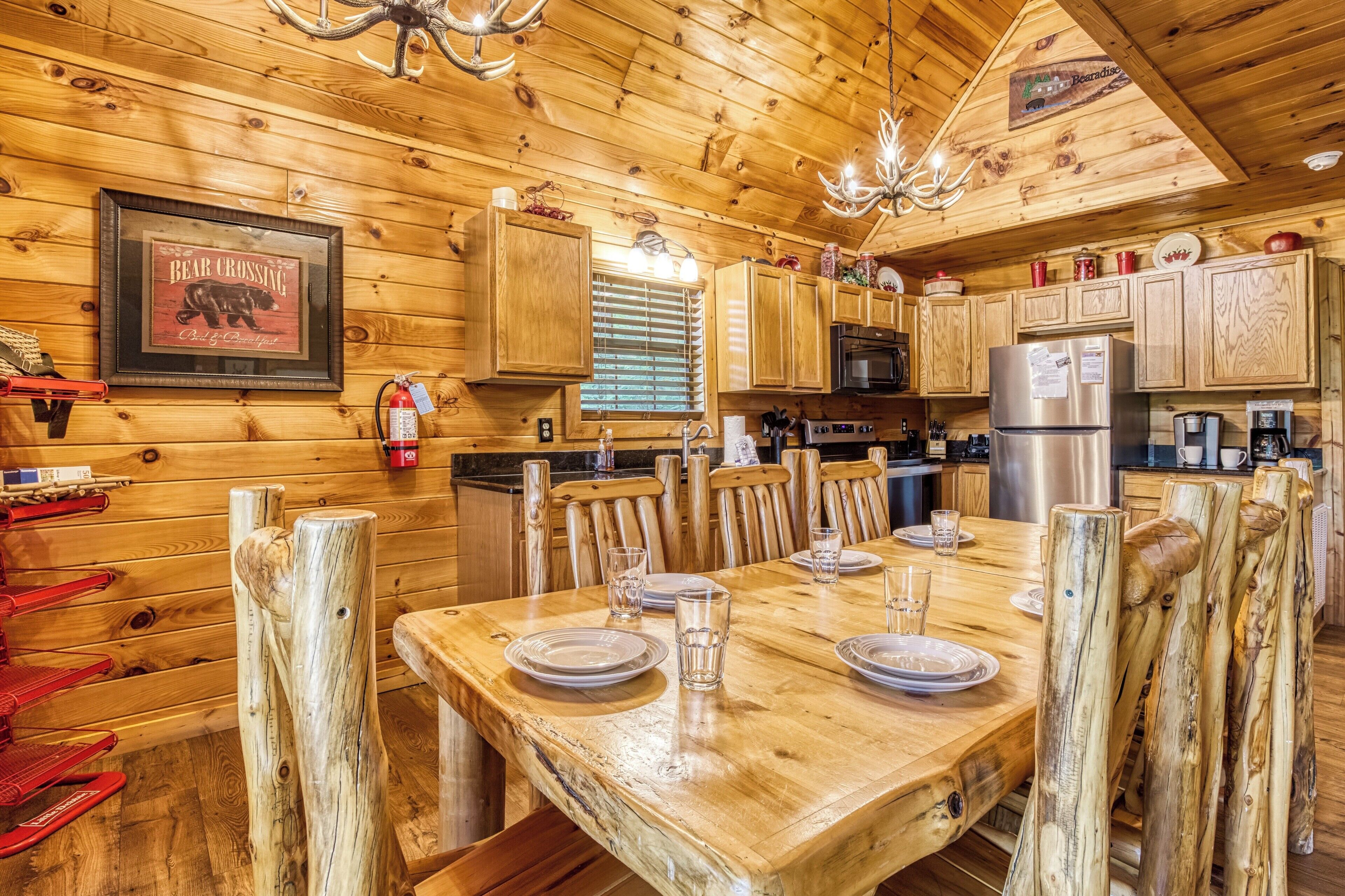 A wooden cabin interior with a dining table set for four, wooden chairs, a kitchen area with a refrigerator, and a framed sign on the wall.