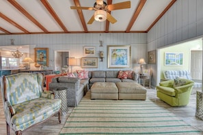 A spacious living room with a high ceiling, wooden beams, and a ceiling fan. There are multiple seating options including a sofa, armchairs, and a green chair. The room features a large window and a view of the outdoors.