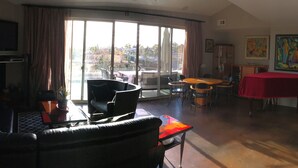 Ein Wohnzimmer mit einer Couch, einem Couchtisch, einem Esstisch mit Stühlen und einem roten Tisch. Ein großes Fenster mit Vorhängen, ein Fernseher und Kunstwerke an den Wänden.