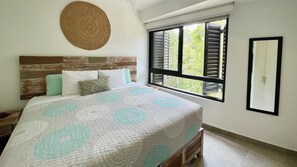 A bedroom with a bed, a window with shutters, a woven wall decoration, and a framed mirror.