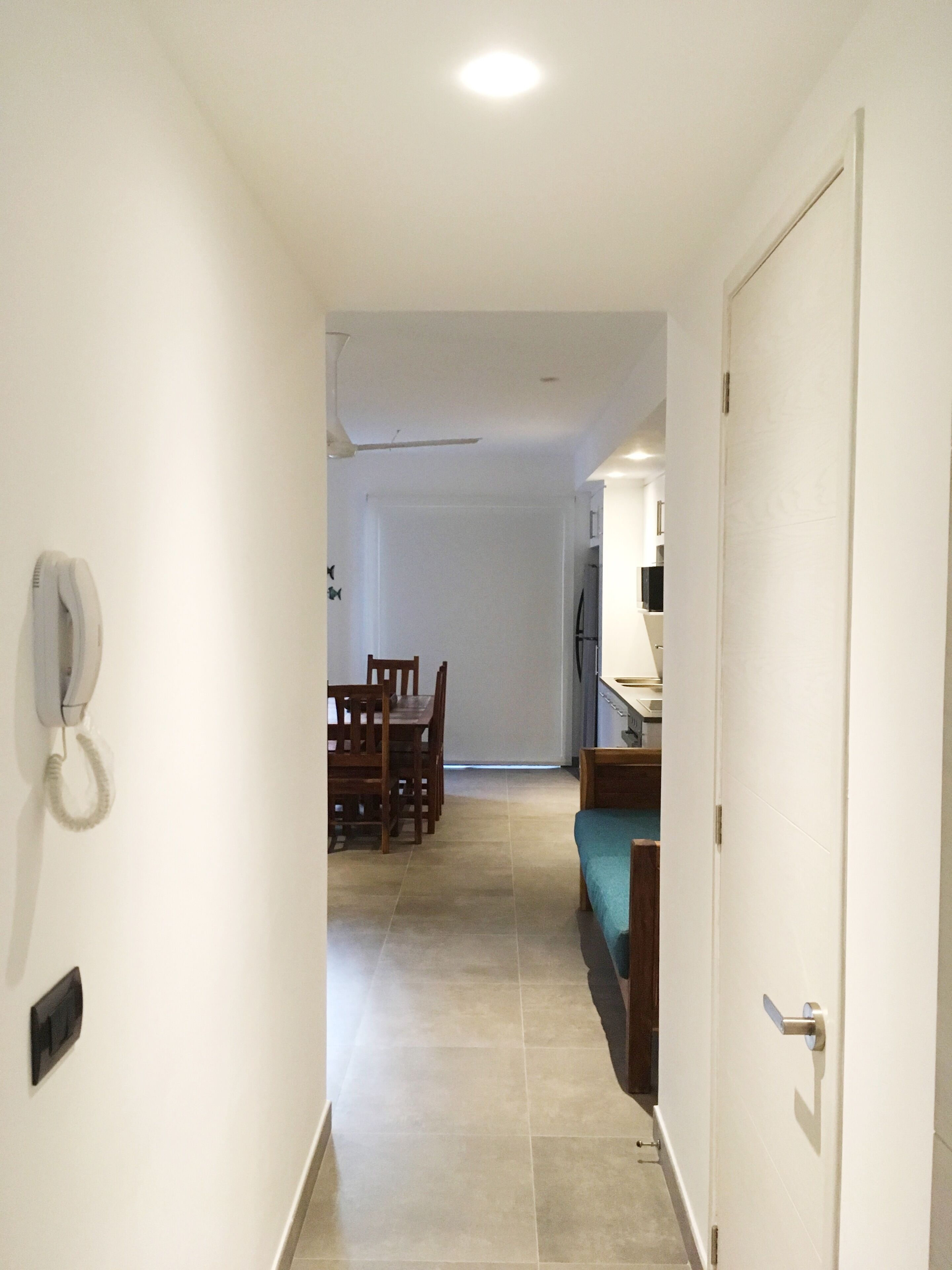 A narrow hallway with a dining area at the end, a door on the right, and a telephone on the left wall.