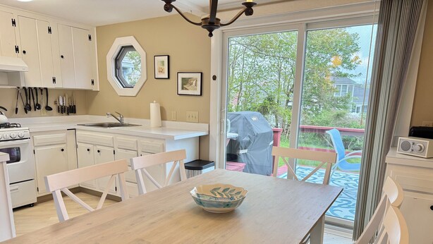 Een keuken met witte kastjes, een eettafel met stoelen, een barbecue buiten, en uitzicht op bomen door het raam.
