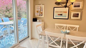 Een zithoek met een tafel en stoelen, een raam met uitzicht op roze bloemen, een wandlamp en ingekaderde foto's.