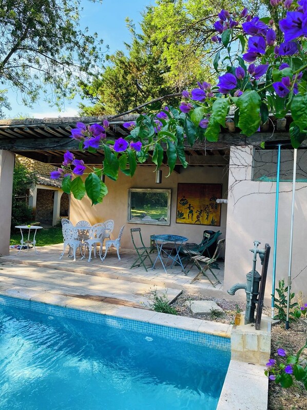 A pool area with a pergola covered in purple flowers, outdoor furniture, and a stone wall with a painting.