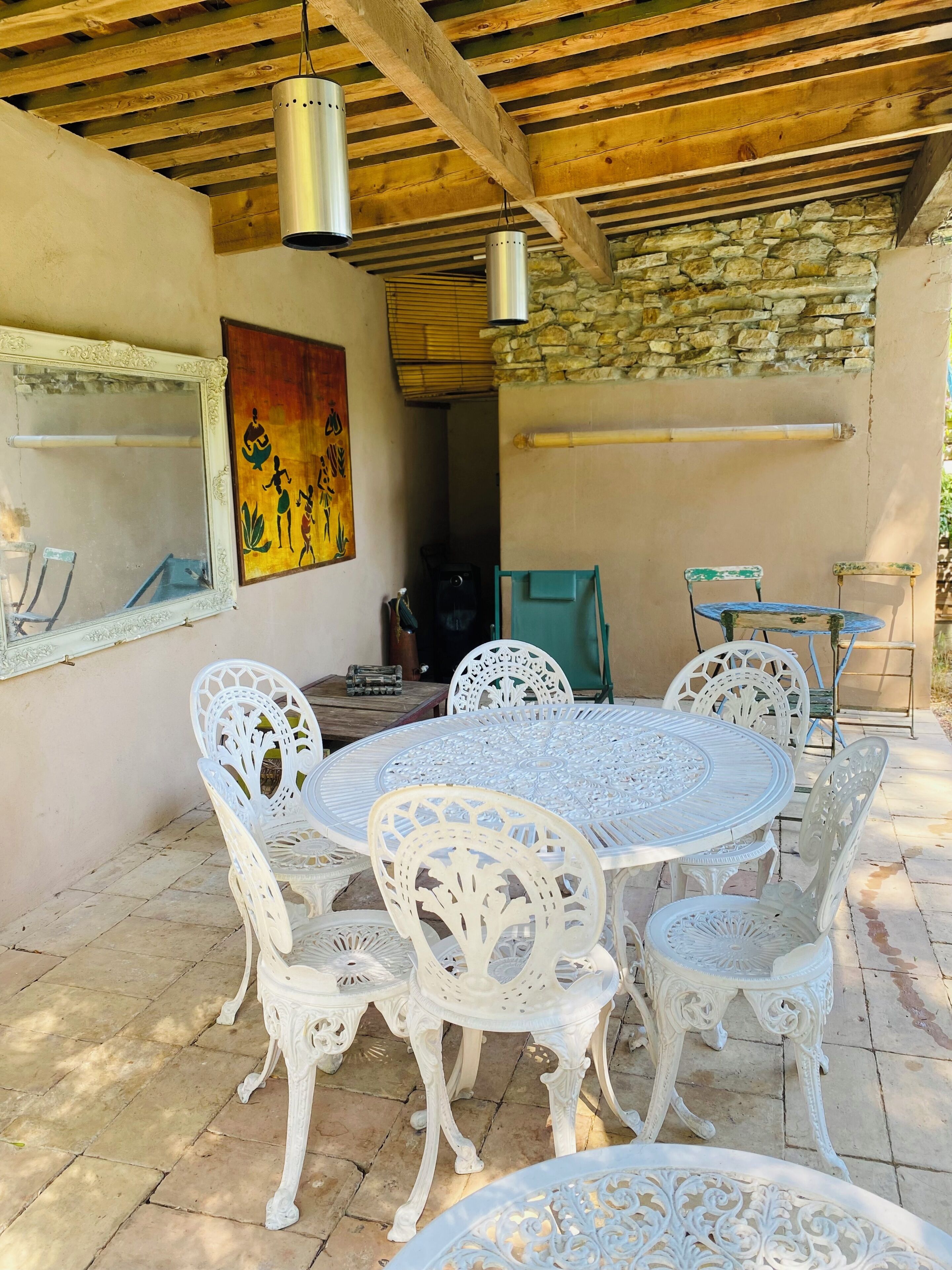 Un patio techado con una mesa redonda y sillas blancas, un muro de piedra y un cuadro en la pared.