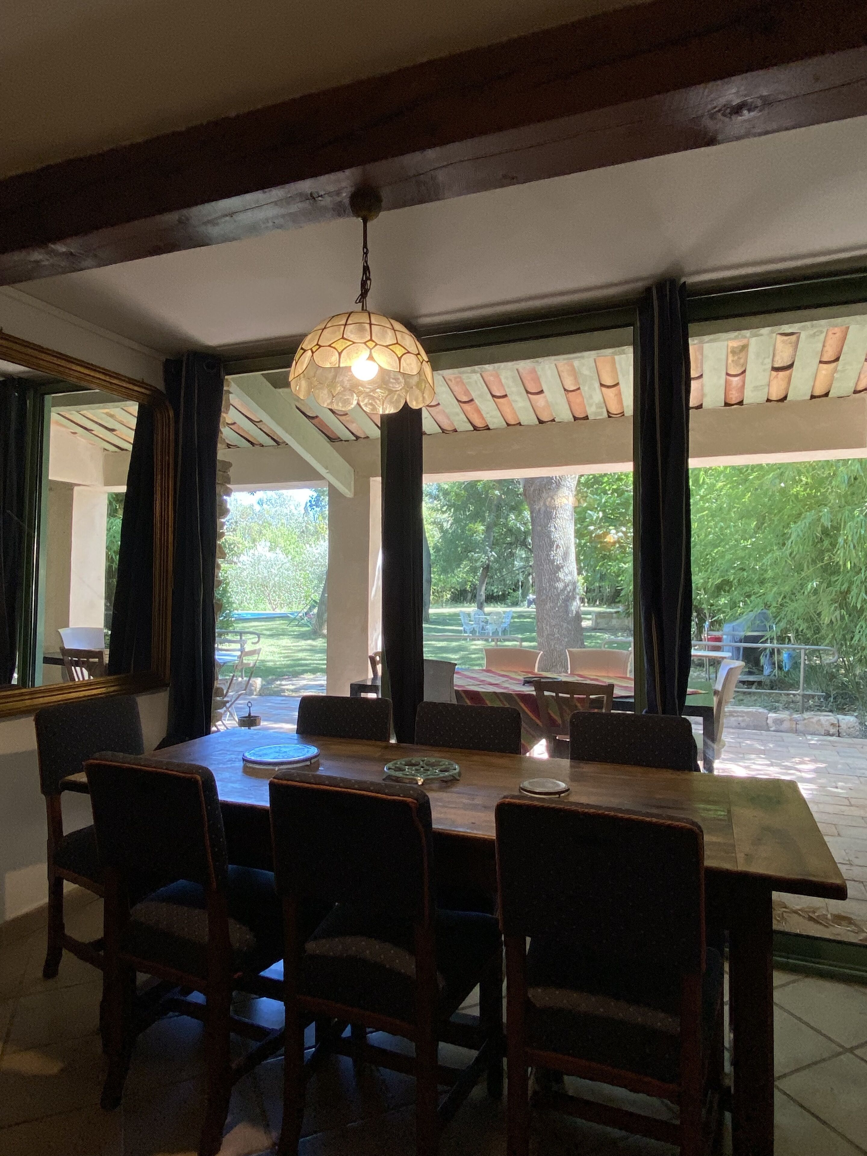 A dining area with a wooden table and chairs, a hanging light fixture, and large windows overlooking a garden.