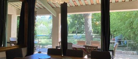 A dining area with a wooden table and chairs, a hanging light fixture, and large windows overlooking a garden.