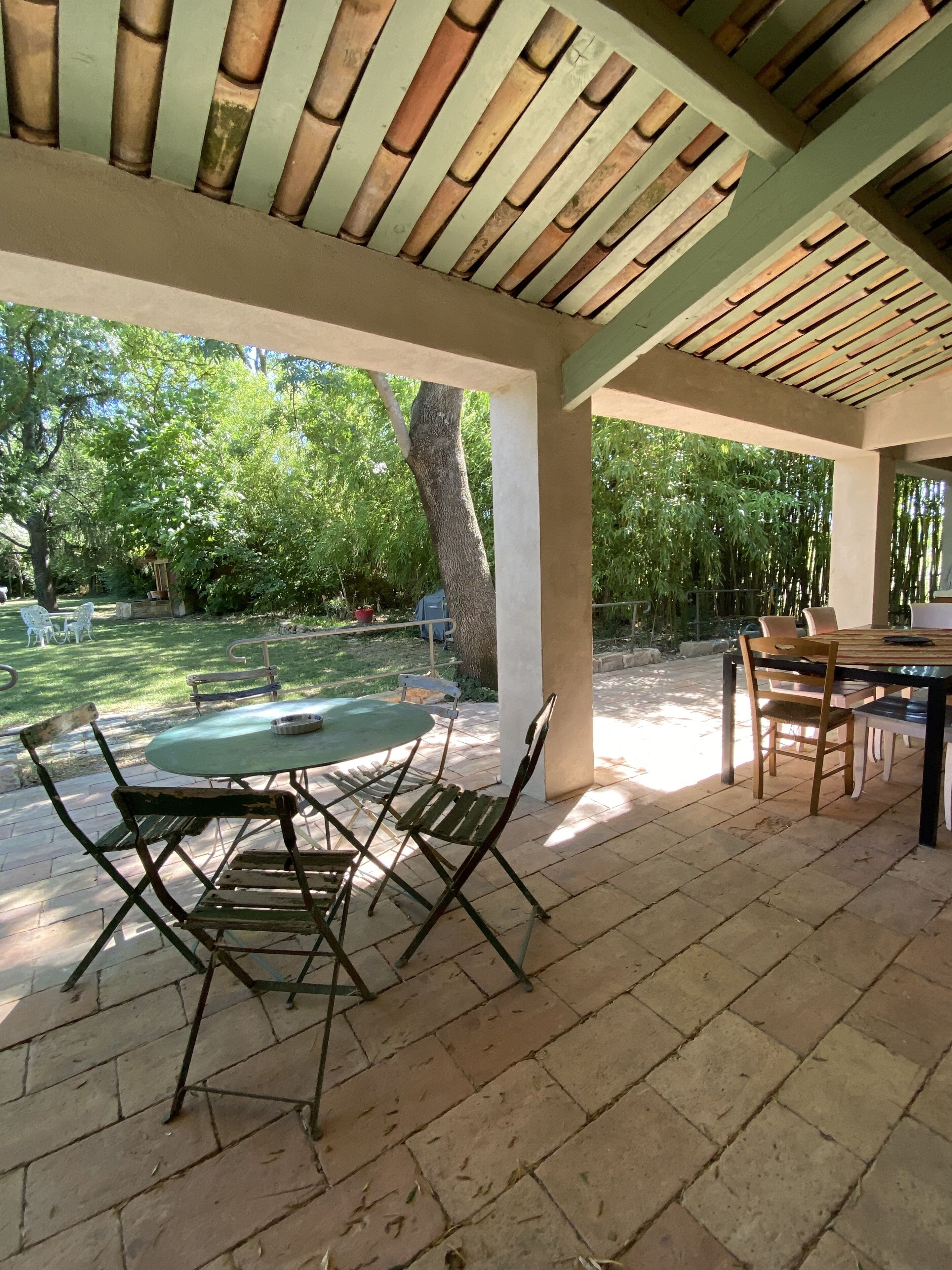 An outdoor patio area with a table and chairs, a covered seating area, and a view of a backyard with trees and a swimming pool.