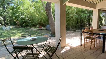 An outdoor patio area with a table and chairs, a covered seating area, and a view of a backyard with trees and a swimming pool.