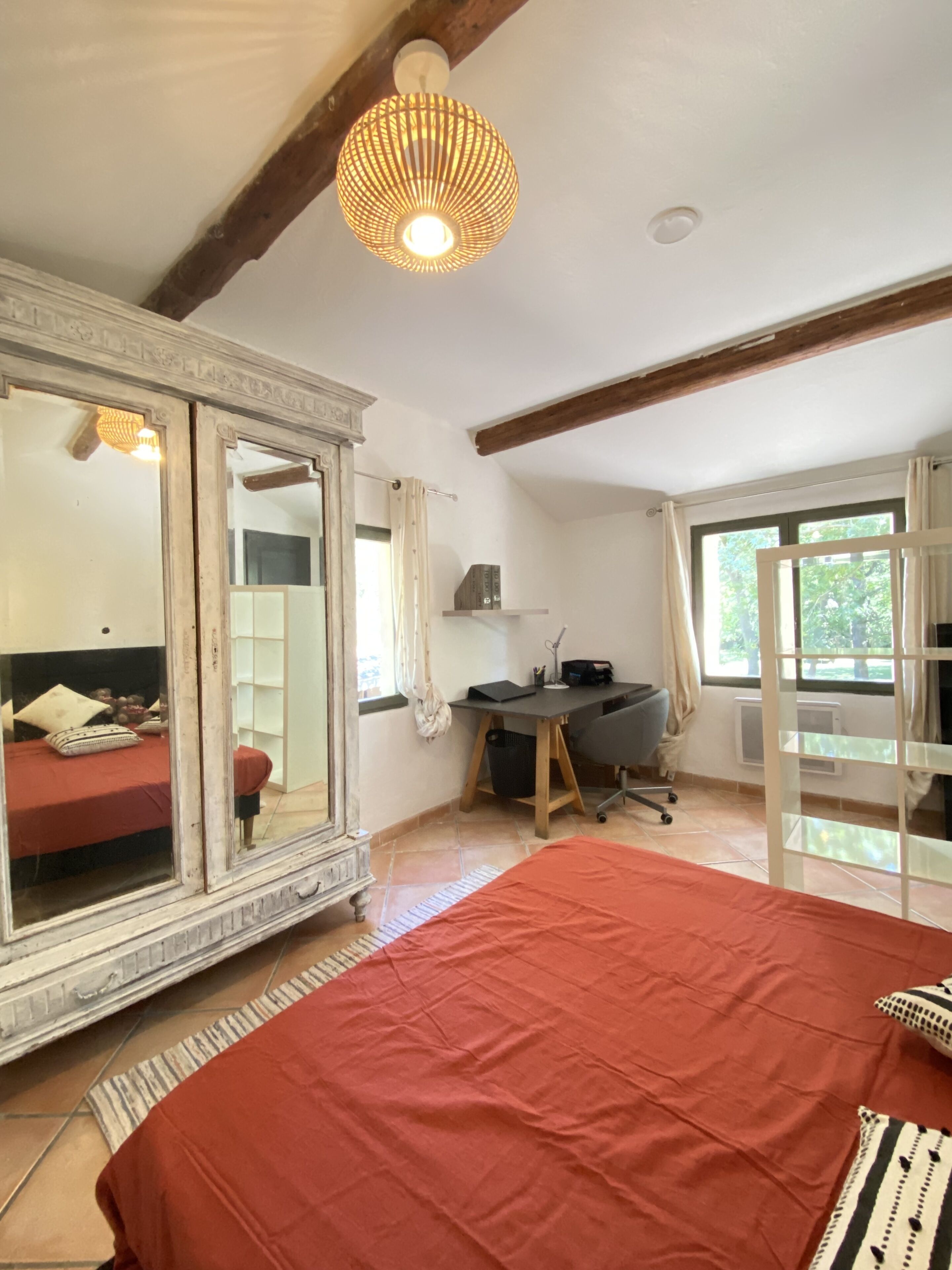 A bedroom with a large bed, a desk, and a chair. There is a mirror, a window, and a ceiling light fixture.