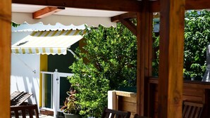 A covered outdoor dining area with wooden tables and chairs, a white door, and a green bush in the background.
