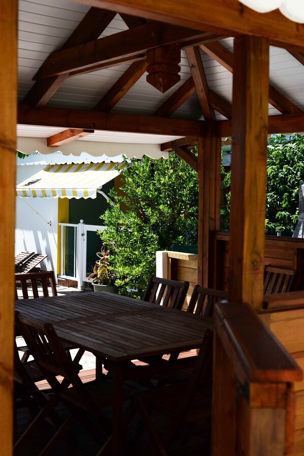 A covered outdoor dining area with wooden tables and chairs, a white door, and a green bush in the background.