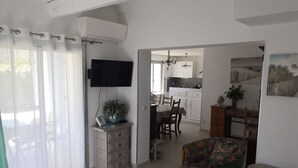 A modern living room with a flat-screen TV, a dining area with a table and chairs, and a kitchen in the background.