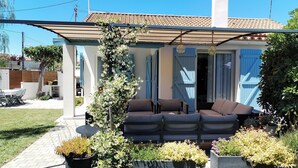 A covered outdoor seating area with a sofa and cushions, surrounded by potted plants and a pergola.