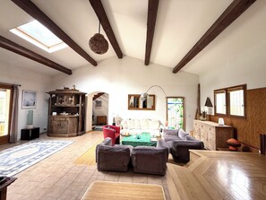 A spacious living room with a high ceiling, wooden beams, a skylight, and a view of the outdoors.