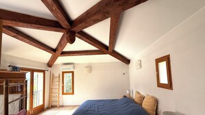 Een slaapkamer met een schuine kap, houten balken, een bed met blauw beddengoed, een nachtkastje met een lamp en een raam met uitzicht op bomen.