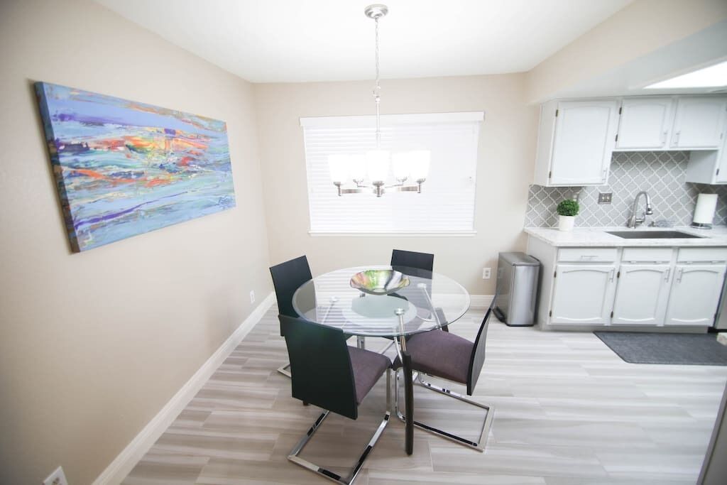 A dining area with a round glass table, four chairs, a chandelier, a window with blinds, and a kitchen area with white cabinets.