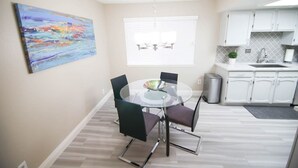 A dining area with a round glass table, four chairs, a chandelier, a window with blinds, and a kitchen area with white cabinets. - Gannet ·- Adorable Condo By Strip- Restaurants,Shopping (Las Vegas)