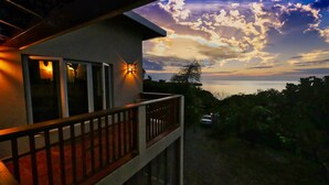 Een balkon met een hekwerk, een verlichte wandlamp en uitzicht op zee bij zonsondergang.