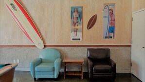 A room with a beige wall, a surfboard, two posters, a blue armchair, a brown leather chair, and a small wooden table. - In the HEART of Ocean Drive in North Myrtle Beach (North Myrtle Beach)