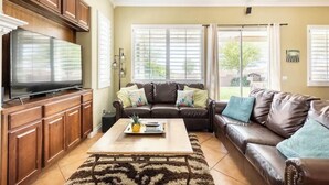 A living room with a brown leather sofa, a coffee table with a tray of items, wooden cabinets, and a flat-screen TV. - Mountain & Sunset Views, Private Salt water Pool Spa, Putting Green, Spacious (Desert Hot Springs)