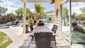 An outdoor patio with a table and chairs, a pergola, and a glass door leading to a pool area. - Mountain & Sunset Views, Private Salt water Pool Spa, Putting Green, Spacious (Desert Hot Springs)