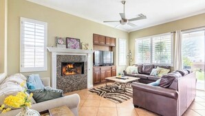 A living room with a fireplace, a ceiling fan, and a large window. - Mountain & Sunset Views, Private Salt water Pool Spa, Putting Green, Spacious (Desert Hot Springs)