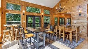 A wooden cabin interior with a large dining table, multiple chairs, and large windows offering views of the surrounding forest.