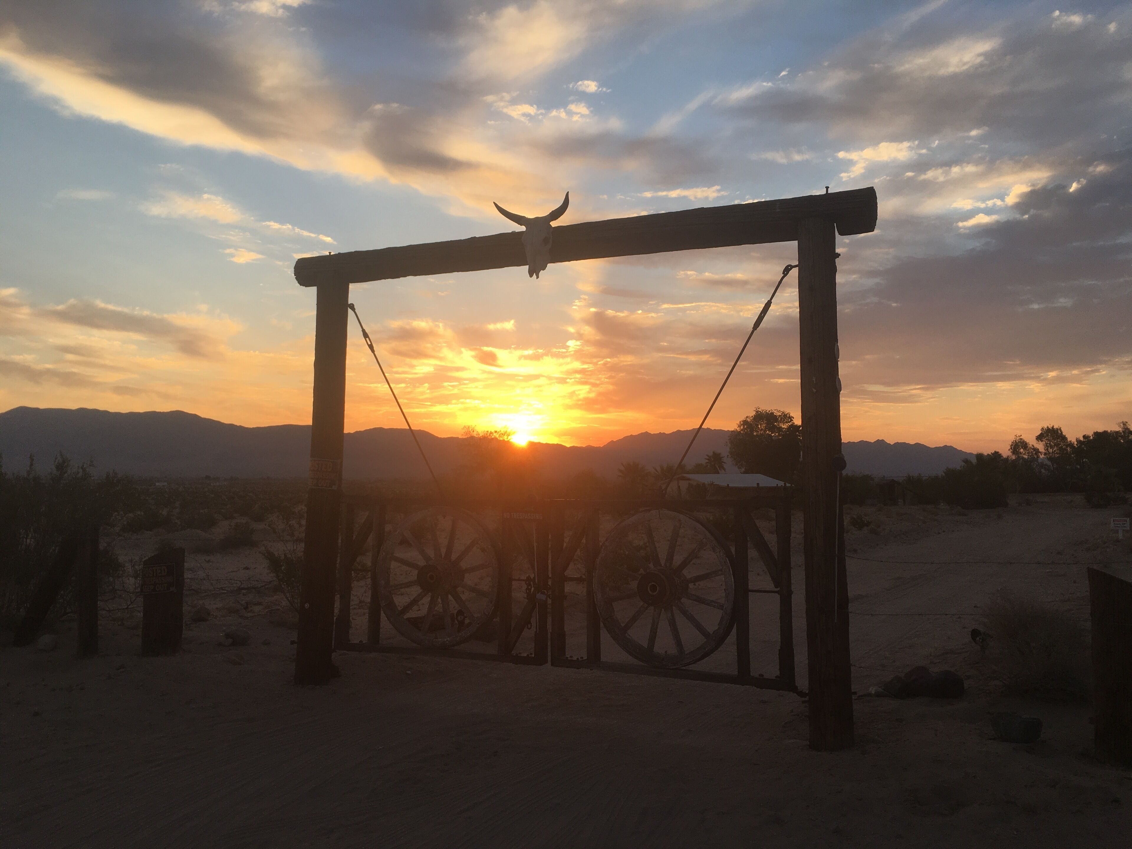A rustic gate with a skull at the top, set against a sunset with mountains in the background.