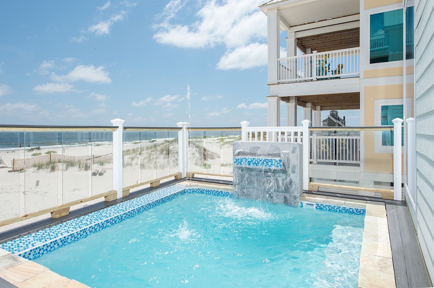 A pool with a waterfall feature, surrounded by a white fence and overlooking a beach.