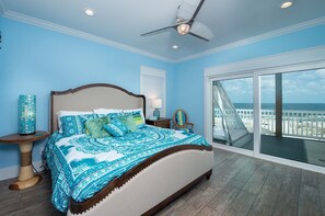 A bedroom with a large bed, a ceiling fan, a nightstand, a lamp, and a view of the ocean through a sliding glass door.