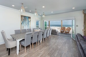 A dining area with a long table and chairs, a large abstract painting, and a view of the ocean through a glass door.