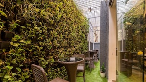 A greenhouse with a glass ceiling, green artificial turf, wicker furniture, and a dense wall of climbing plants. - Stylotel By Jagadish (Bengaluru)