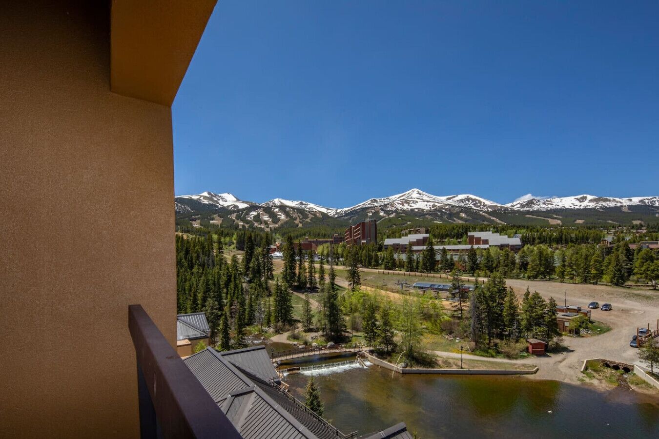 A view from a balcony overlooking a mountainous landscape with snow-capped peaks, a small lake, and a parking area with buildings in the background.